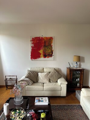 Uma sala de estar com um sofá, uma mesa de centro com diversos objetos, uma estante e um quadro na parede.