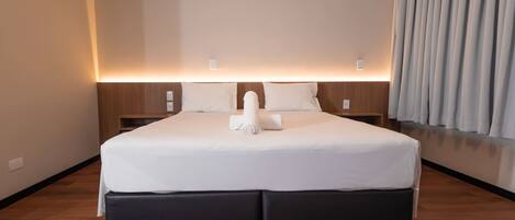 Habitación de hotel con una cama grande, cabecera con iluminación integrada, mesita de noche de madera y ventana con cortinas.