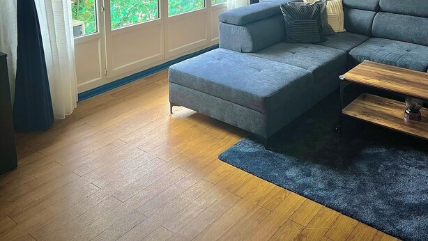 Una sala de estar con un sofá seccional gris, una mesa de centro de madera y un tapete azul. Hay ventanas grandes con cortinas, un jarrón con plantas secas y obras de arte enmarcadas en la pared.