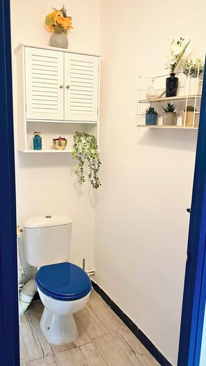 A small bathroom with a white toilet, a blue seat, a white cabinet with horizontal lines, and a shelf with plants and a vase. - Blue Seahorse Flat - T3 Avignon Intra-muros - GARAGE WIFI CLIM (Avignon)