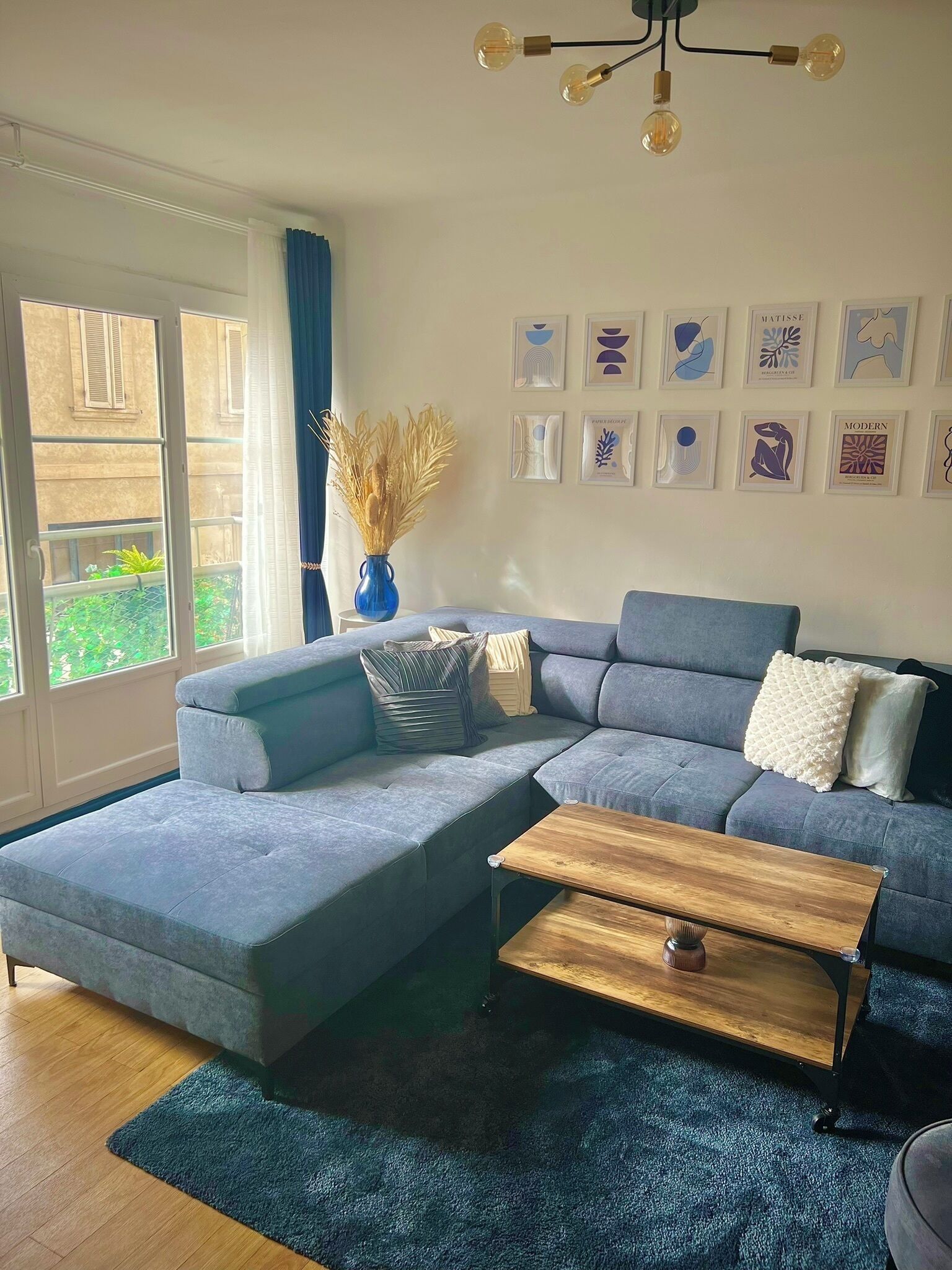 Ein modernes Wohnzimmer mit einem grauen Ecksofa, einem Holztisch und einem blauen Teppich. An der Wand hängen gerahmte Kunstwerke und auf einem Beistelltisch steht eine Vase mit Trockenpflanzen.