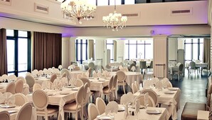 Un gran saló de banquets amb diverses taules rodones, cadires blanques i lluminàries penjants.