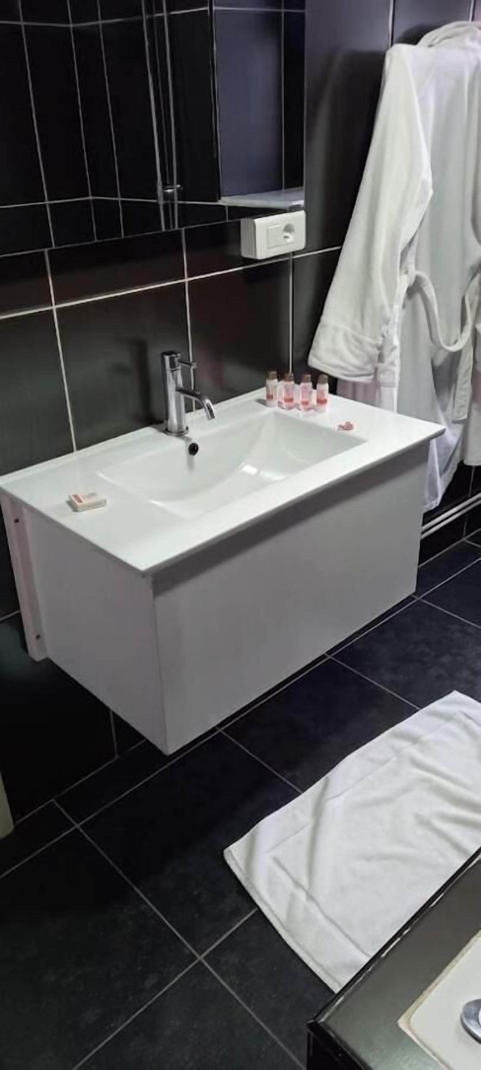 A modern bathroom with a white sink, black tiles, and a white towel on the floor.