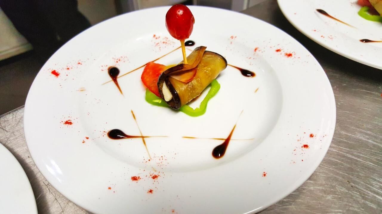 Un plato blanco con un postre que incluye una porción de pastel, una cereza y salsa cubriendo.
