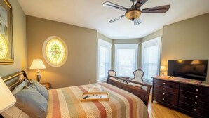 A bedroom with a bed, a ceiling fan, a TV, a dresser, and a window with blinds. - THE VICTORIAN GUESTHOUSE BED & BREAKFAST - Room 3 (Delphos)
