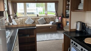 Une cuisine compacte équipée d’une cuisinière, d’un four et d’un évier. On y trouve une fenêtre avec des rideaux, un banc avec des coussins et une vue sur une maison à l’extérieur.