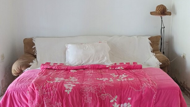 A bed with a pink floral bedspread, a white pillow, and a brown pillow. - Green slice (Mairinque)