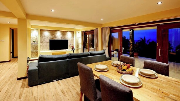 Un comedor moderno con una mesa de madera preparada para cuatro personas, un televisor de pantalla plana grande montado en la pared y un gran ventanal corredero que da a un espacio al aire libre.
