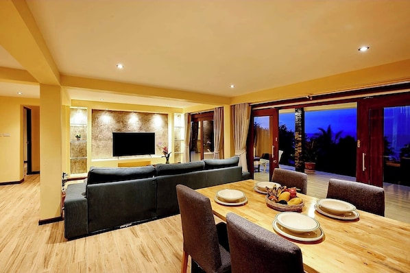 Un comedor moderno con una mesa de madera preparada para cuatro personas, un televisor de pantalla plana grande montado en la pared y un gran ventanal corredero que da a un espacio al aire libre.