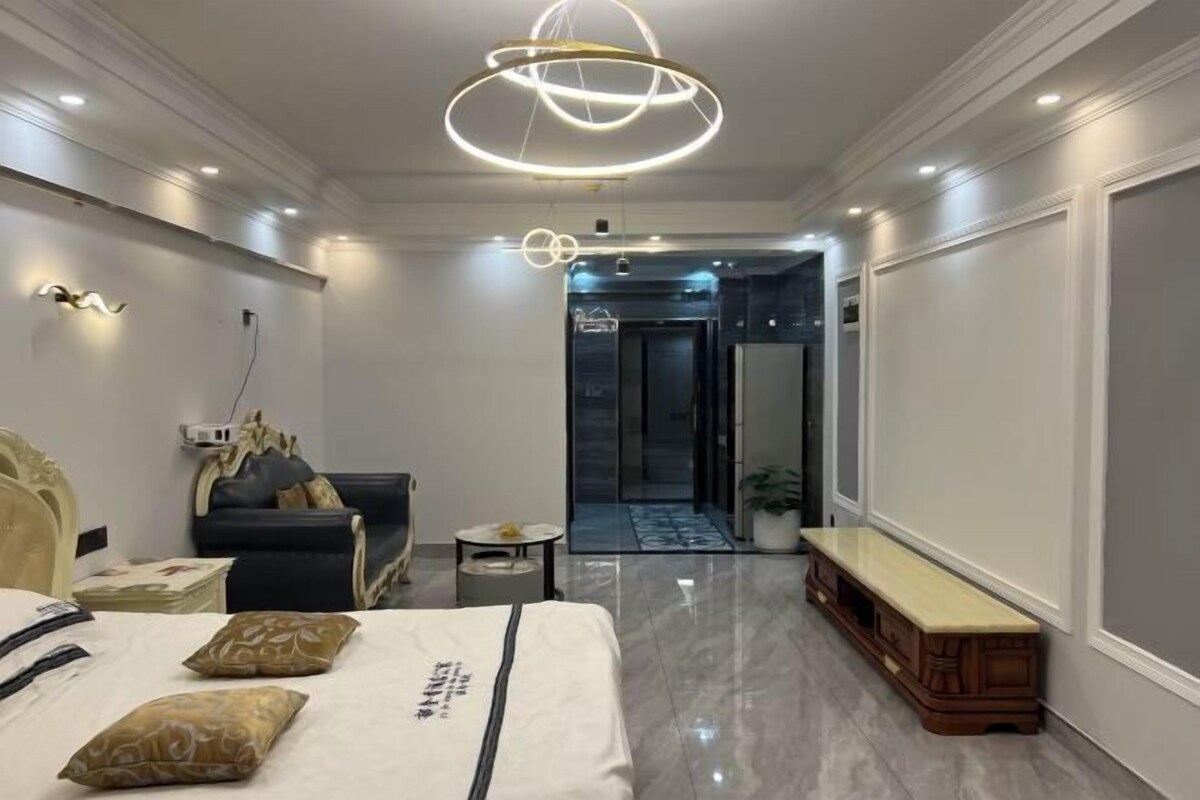 A modern hotel room with a bed, a seating area, and a hallway leading to a door.