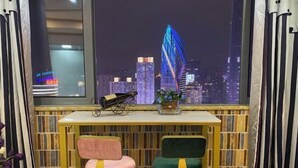 Une pièce avec vue sur les toits d’une ville illuminée la nuit, une petite table avec un télescope et deux chaises.