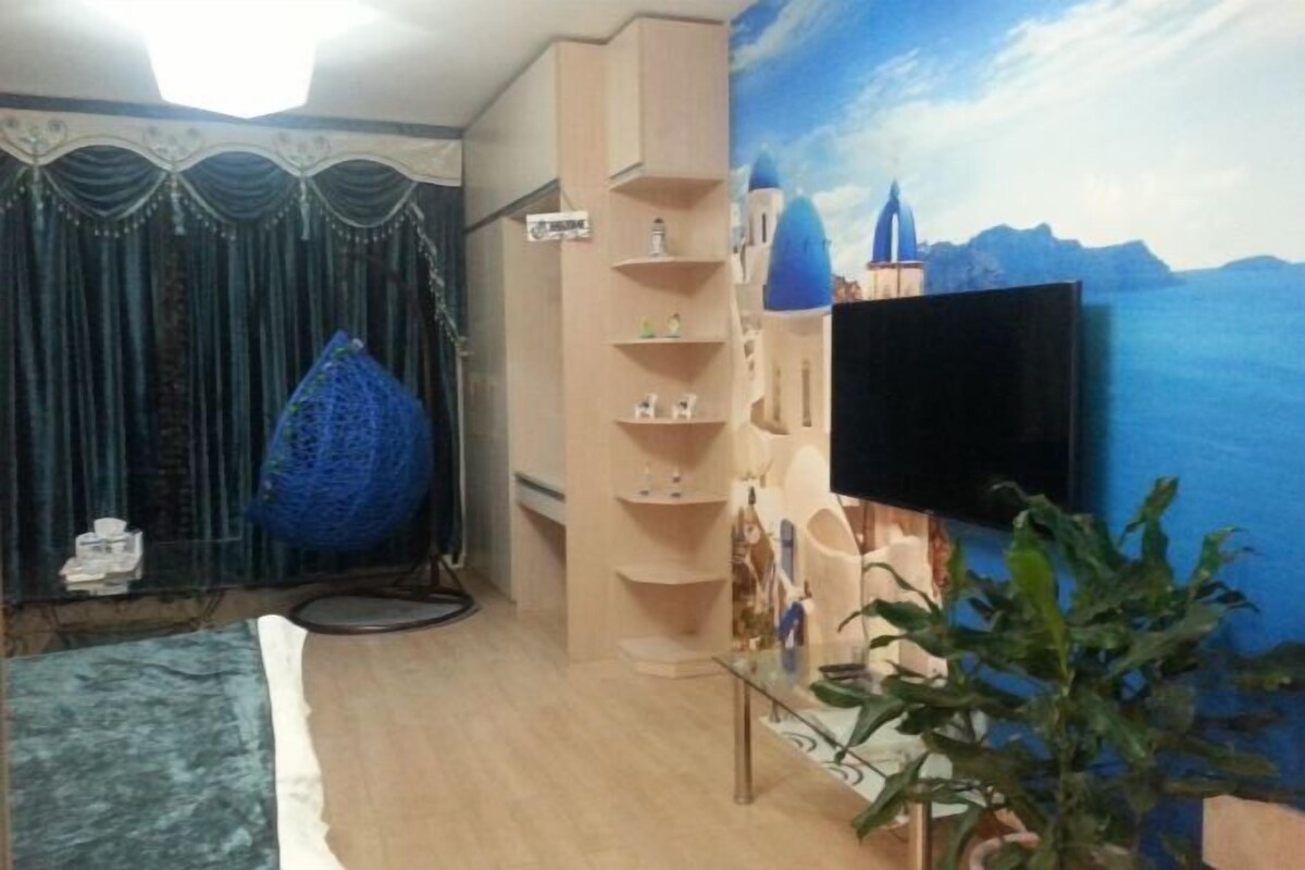 A room with a large TV, a blue hanging decoration, a shelf with items, and a painting on the wall.