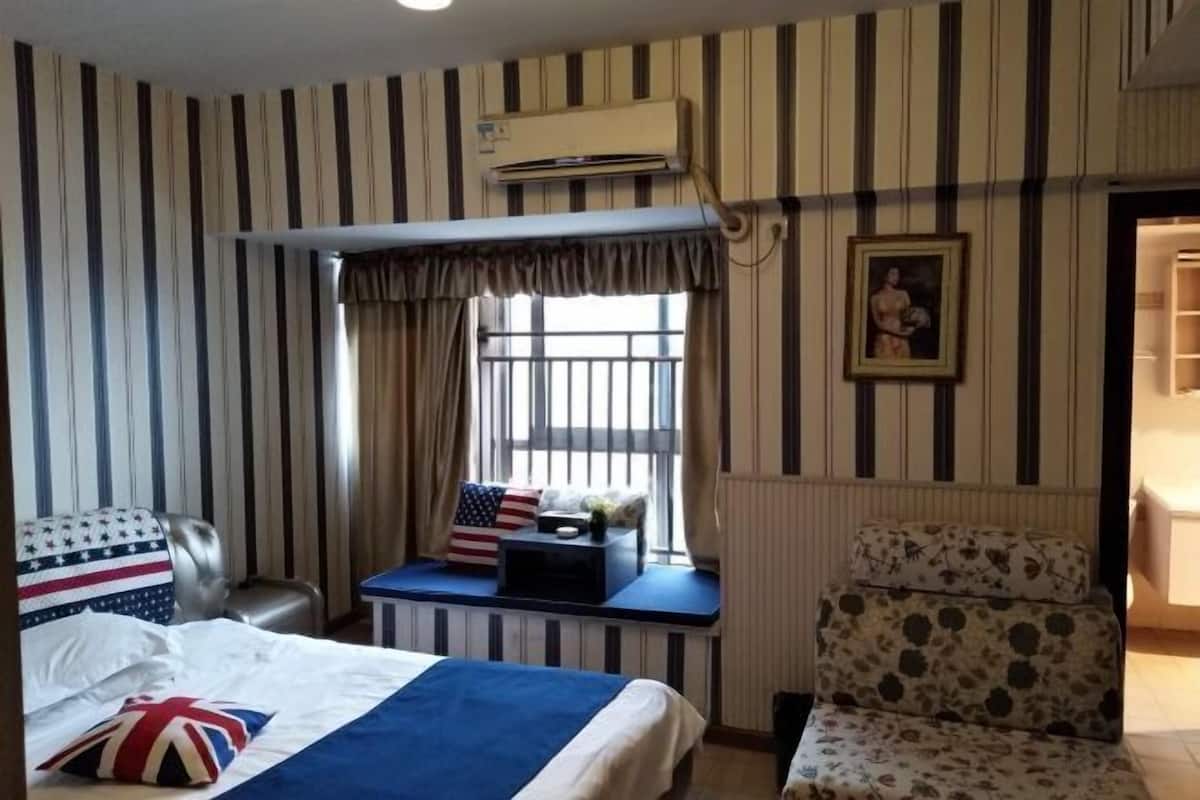A hotel room with a striped wall, a bed with a blue and white bedspread, a seating area with a view, and an air conditioning unit.