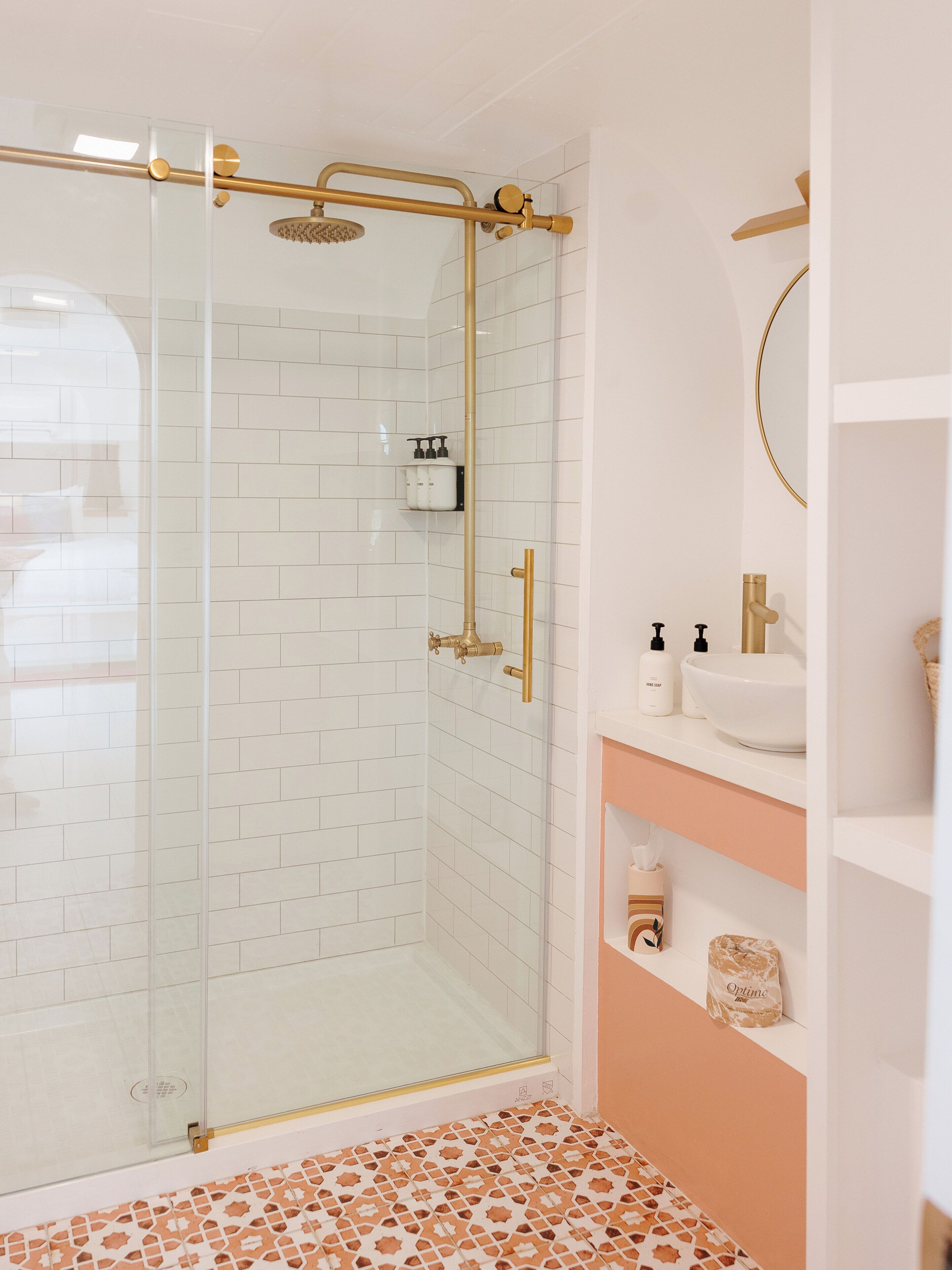 A modern bathroom with a glass-enclosed shower, white tiled walls, and a white sink with a gold faucet.