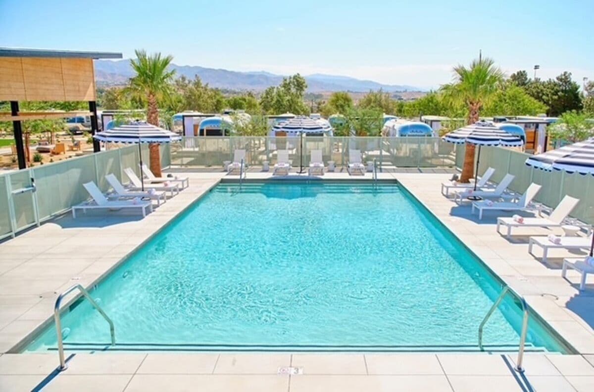A rectangular pool with a clear blue water surface, surrounded by sun loungers and umbrellas, with a view of mountains in the background.