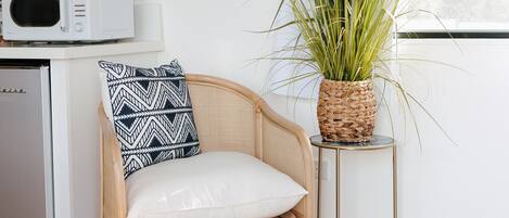 Una silla de mimbre con un cojín blanco y un almohadón estampado, una maceta trenzada y un microondas blanco sobre un mueble blanco.