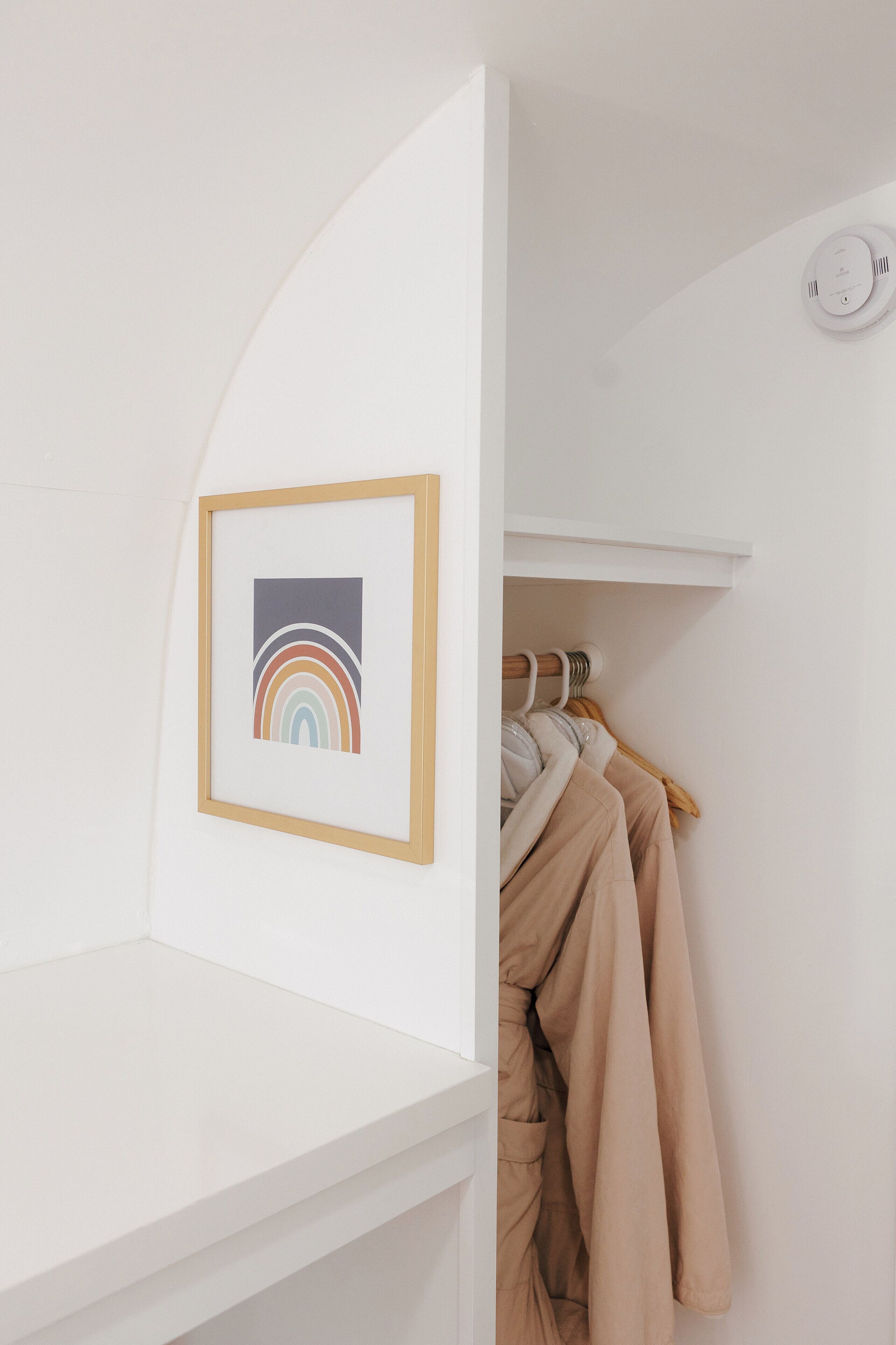 A white room with a framed picture of a rainbow, a coat rack with clothes, and a smoke detector on the ceiling.