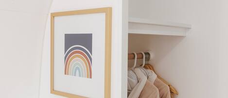 A white room with a framed picture of a rainbow, a coat rack with clothes, and a smoke detector on the ceiling.