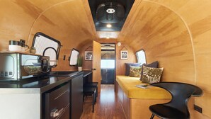 A compact kitchen with a stove, microwave, and coffee maker. There is a bench and a small table with a plant. The room has wooden walls and a curved ceiling. - Acres Landing (Desert Hot Springs)