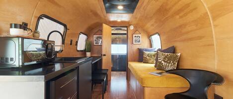 A compact kitchen with a stove, microwave, and coffee maker. There is a bench and a small table with a plant. The room has wooden walls and a curved ceiling.