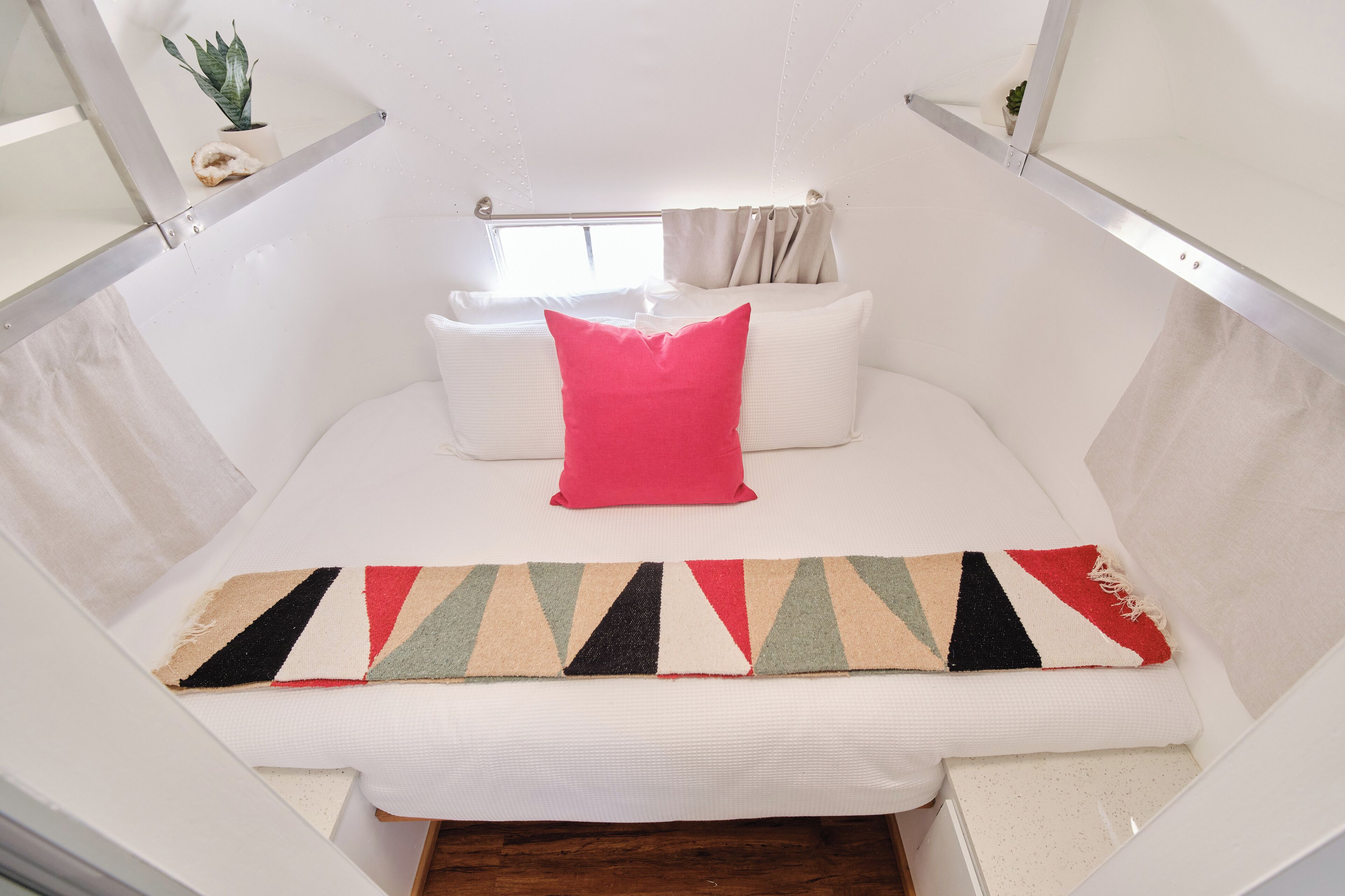 Un letto con materasso bianco, un cuscino rosso e una coperta a righe. Si vede una finestra con tende e una piccola pianta su uno scaffale sopra il letto.