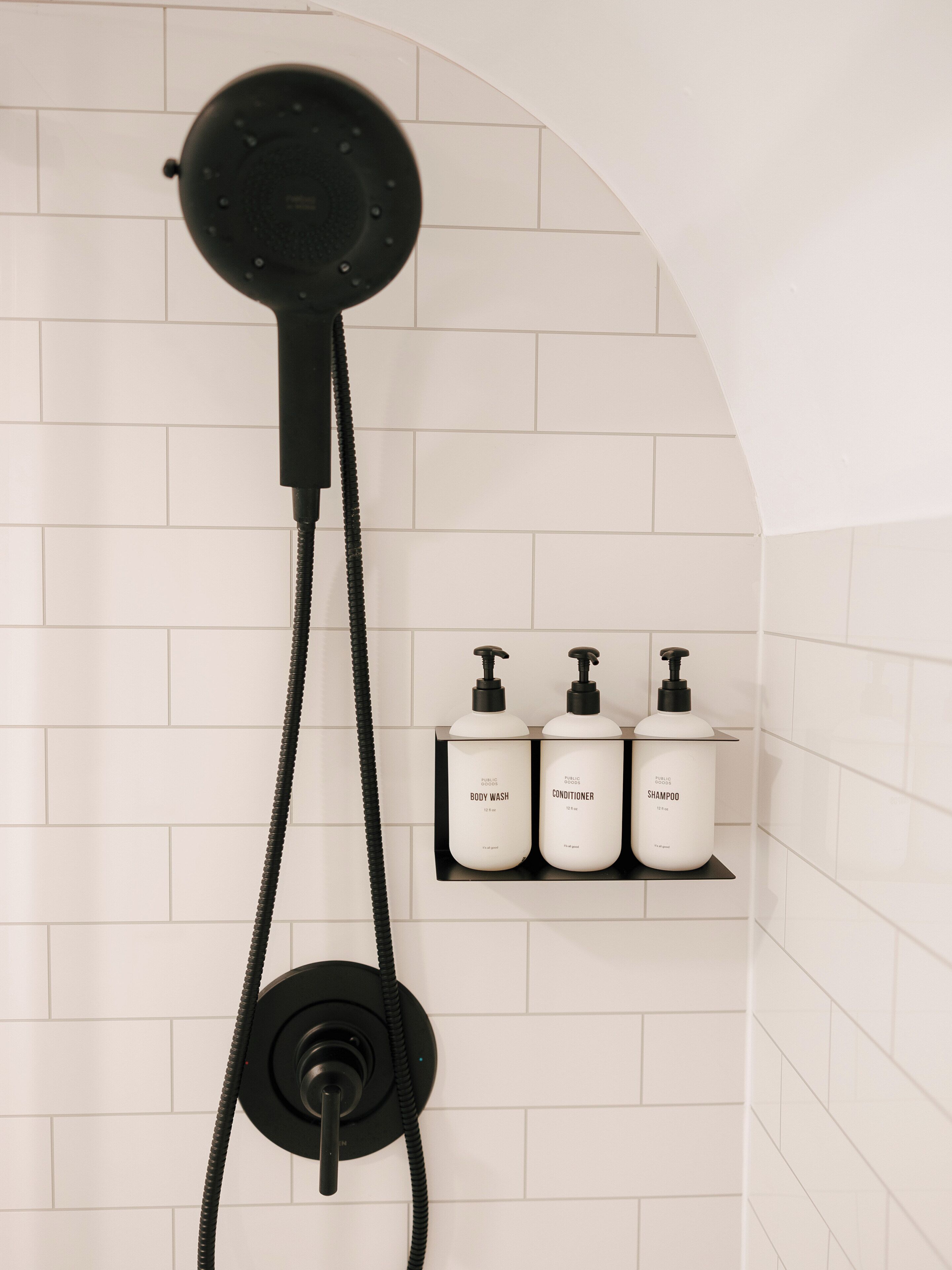 A shower with a black hose and a wall-mounted control. There are three white bottles on a black shelf labeled 'BODY WASH,' 'CONDITIONER,' and 'SHAMPOO.'