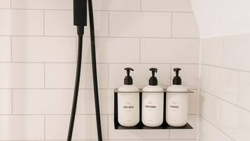 A shower with a black hose and a wall-mounted control. There are three white bottles on a black shelf labeled 'BODY WASH,' 'CONDITIONER,' and 'SHAMPOO.'