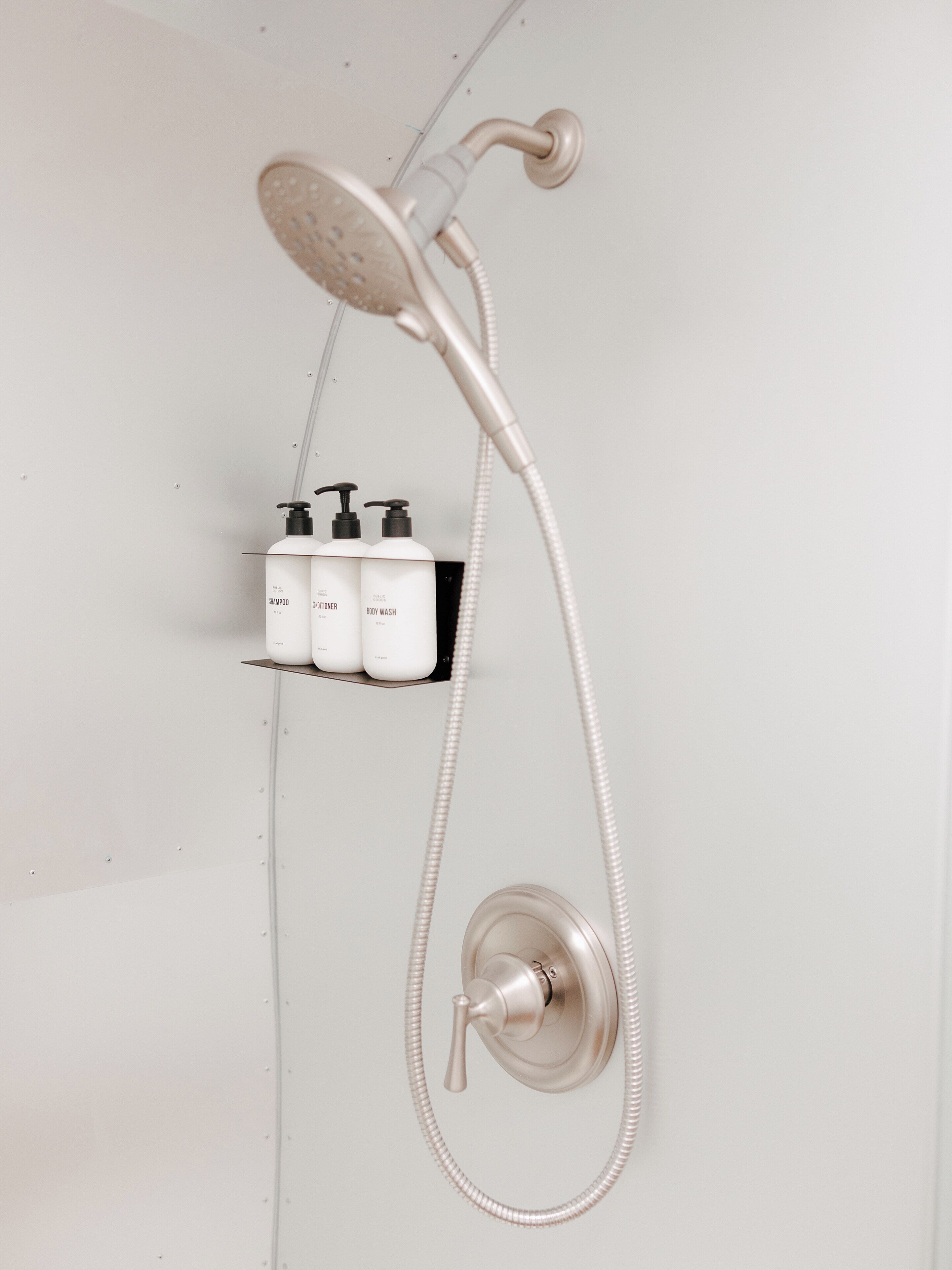 A shower with a handheld showerhead and a wall-mounted control. There are three bottles of shampoo, conditioner, and body wash on a shelf below.