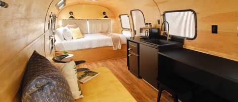 A compact, well-lit interior with a bed, a small kitchenette, and a desk area.