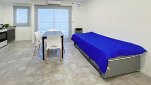 Une petite pièce lumineuse, avec une table et des chaises, un lit recouvert d’une couverture bleue, un coin cuisine et une grande fenêtre donnant sur un paysage agréable.