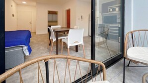 Une chambre moderne comprenant un lit, une table à manger avec des chaises, un fauteuil en osier et une porte vitrée donnant sur un coin cuisine.