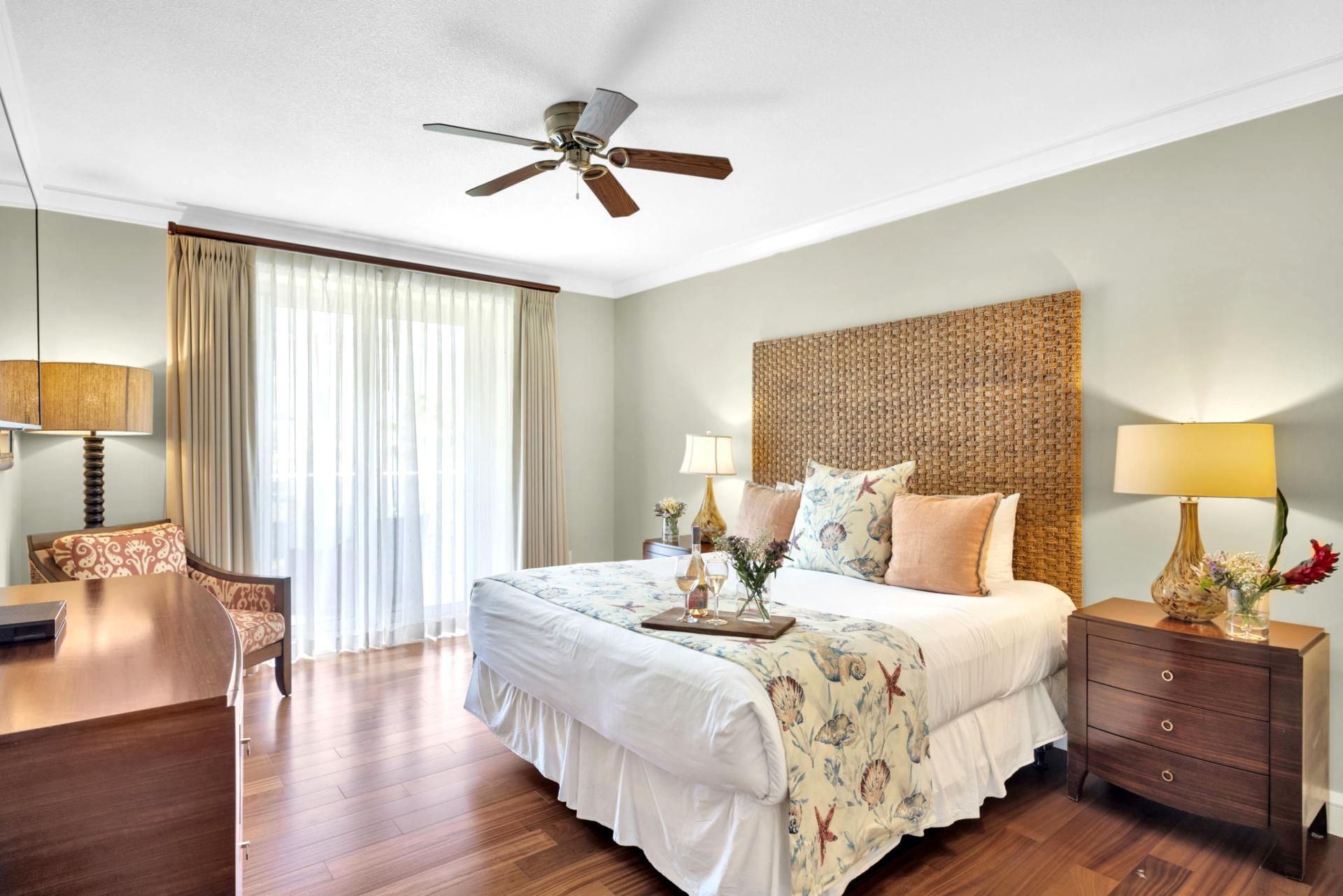 A hotel room with a bed, bedside tables, a desk, a chair, a ceiling fan, and a window with curtains.