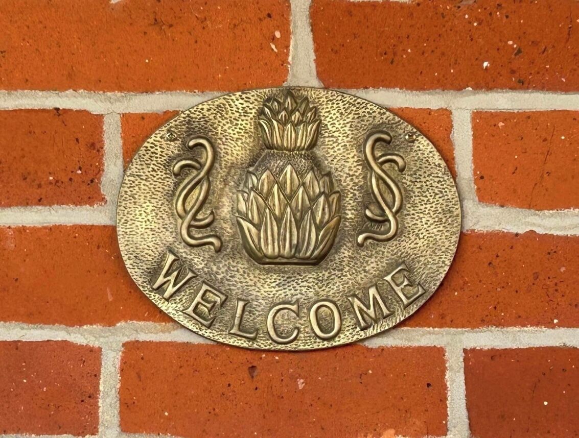 A brass plaque with a pineapple emblem and the words "WELCOME" on a brick wall.
