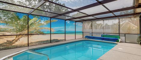 An indoor swimming pool with a view of a beach and palm trees through large windows.