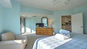 A bedroom with a bed, a TV on a dresser, a chair, and a view of the kitchen through an open door. - Exceptional location!  Across the street from the beach! Walk to Everything!  (Hilton Head Island)