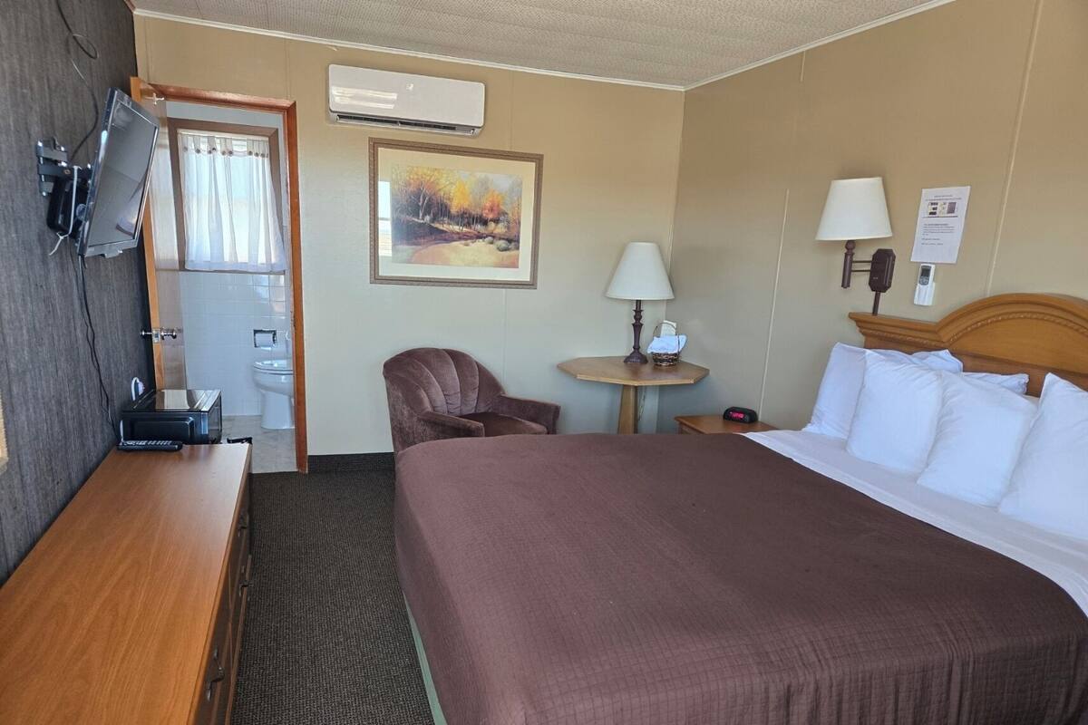 Ein Hotelzimmer mit einem Bett, einem Schreibtisch, einem Sessel, einem kleinen Tisch mit Lampe und einer Klimaanlage.