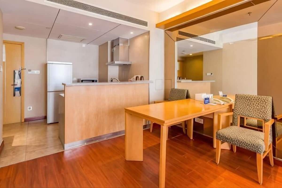 A hotel room with a wooden dining table, a kitchen area with a refrigerator and stove, and a seating area with chairs.