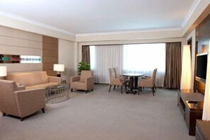 Habitación de hotel con sofá, mesa de comedor con sillas, televisor y ventana grande con cortinas.