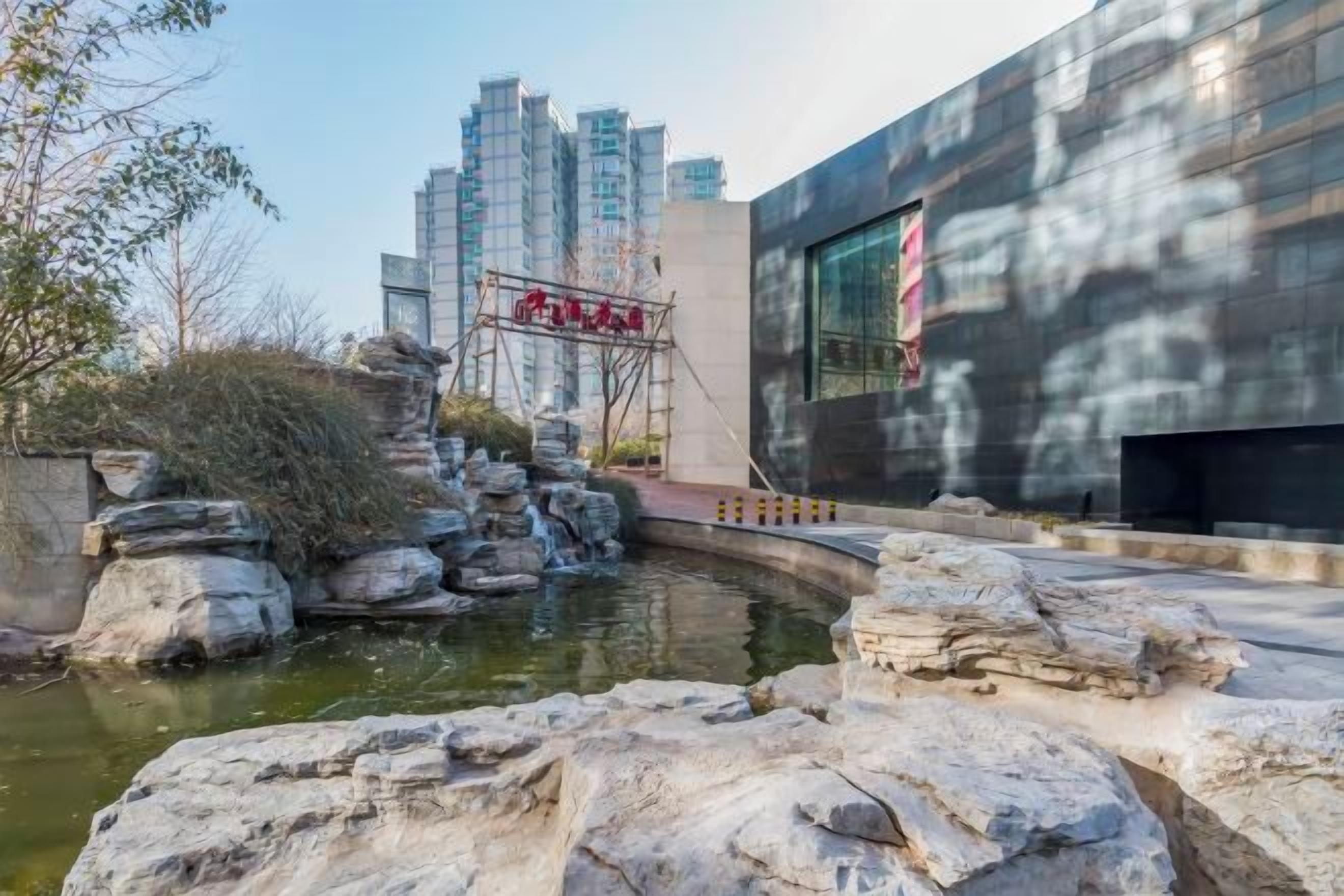 Un edificio moderno con una amplia fachada de vidrio, una fuente rocosa en primer plano y un paisaje urbano al fondo.