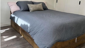 A bedroom with a wooden bed frame, grey bedding, and a window with blinds.