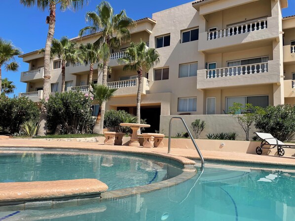 A pool area with a clear blue pool, palm trees, and a building with balconies. - Corona del Mar by Rocky Host (Puerto Peñasco)