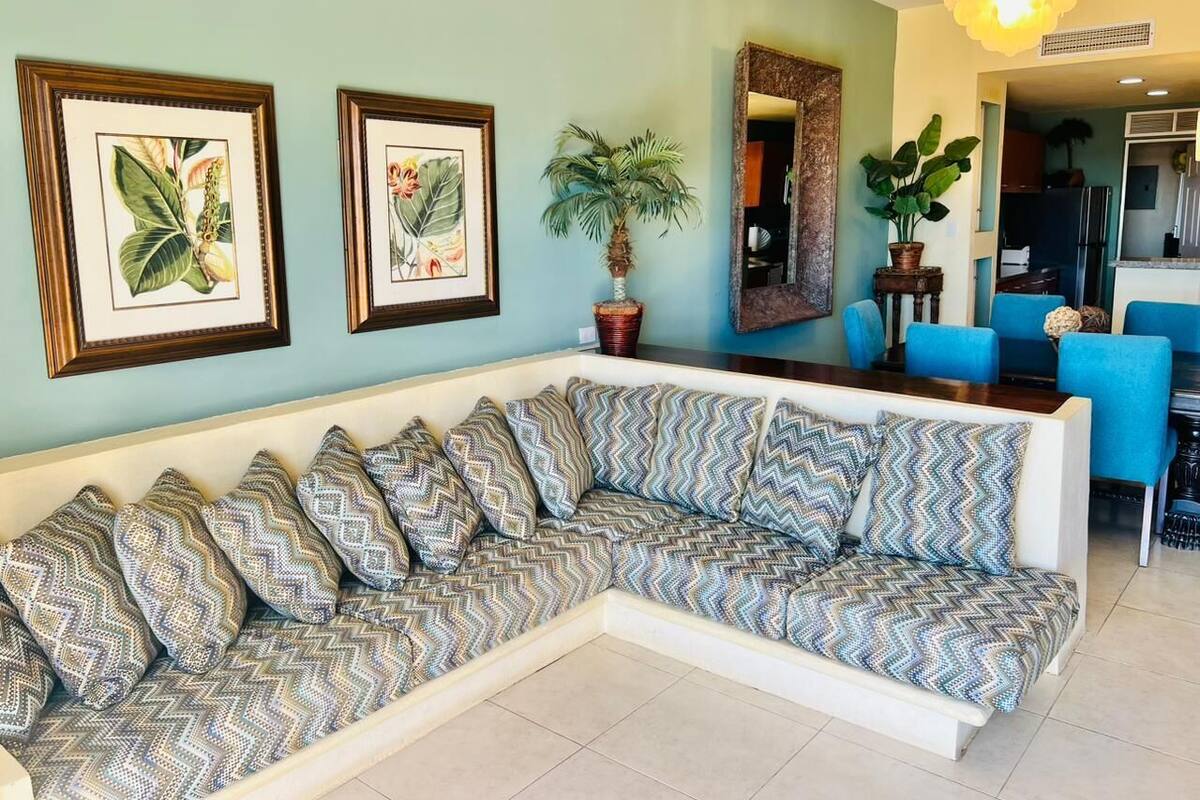 A living room with a large L-shaped sectional sofa, framed botanical prints on the wall, and a dining area with blue chairs.