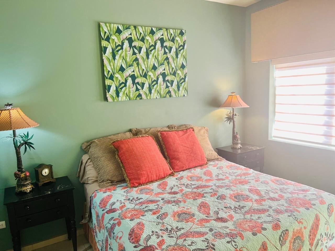 A bedroom with a bed, two nightstands, a wall clock, a lamp, and a window with blinds.