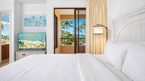 A hotel room with a bed, a television, a painting, a window with a view of palm trees, and a balcony.