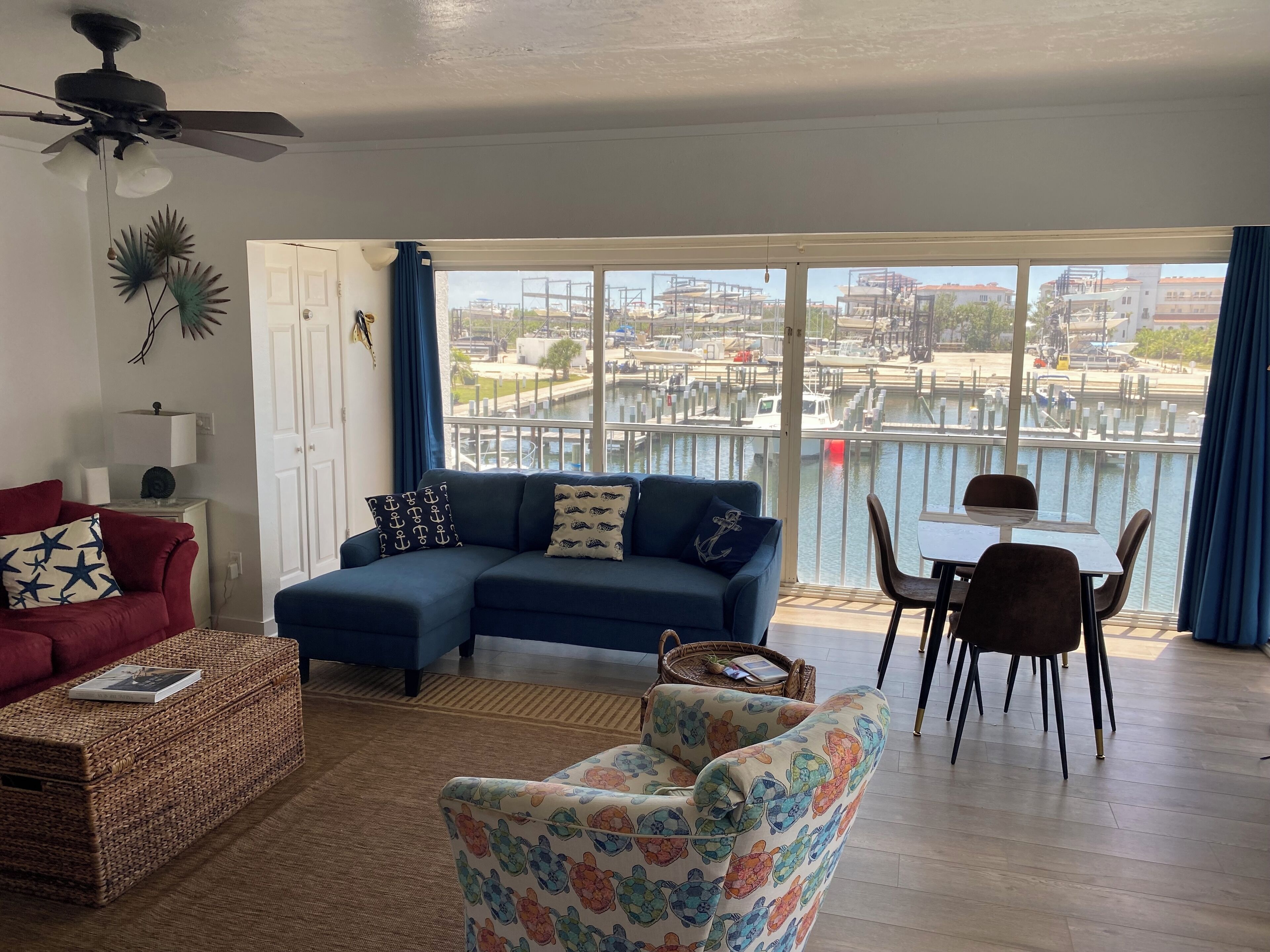 A living room with a large window overlooking a marina, a blue sofa, a red armchair, and a small table with chairs.