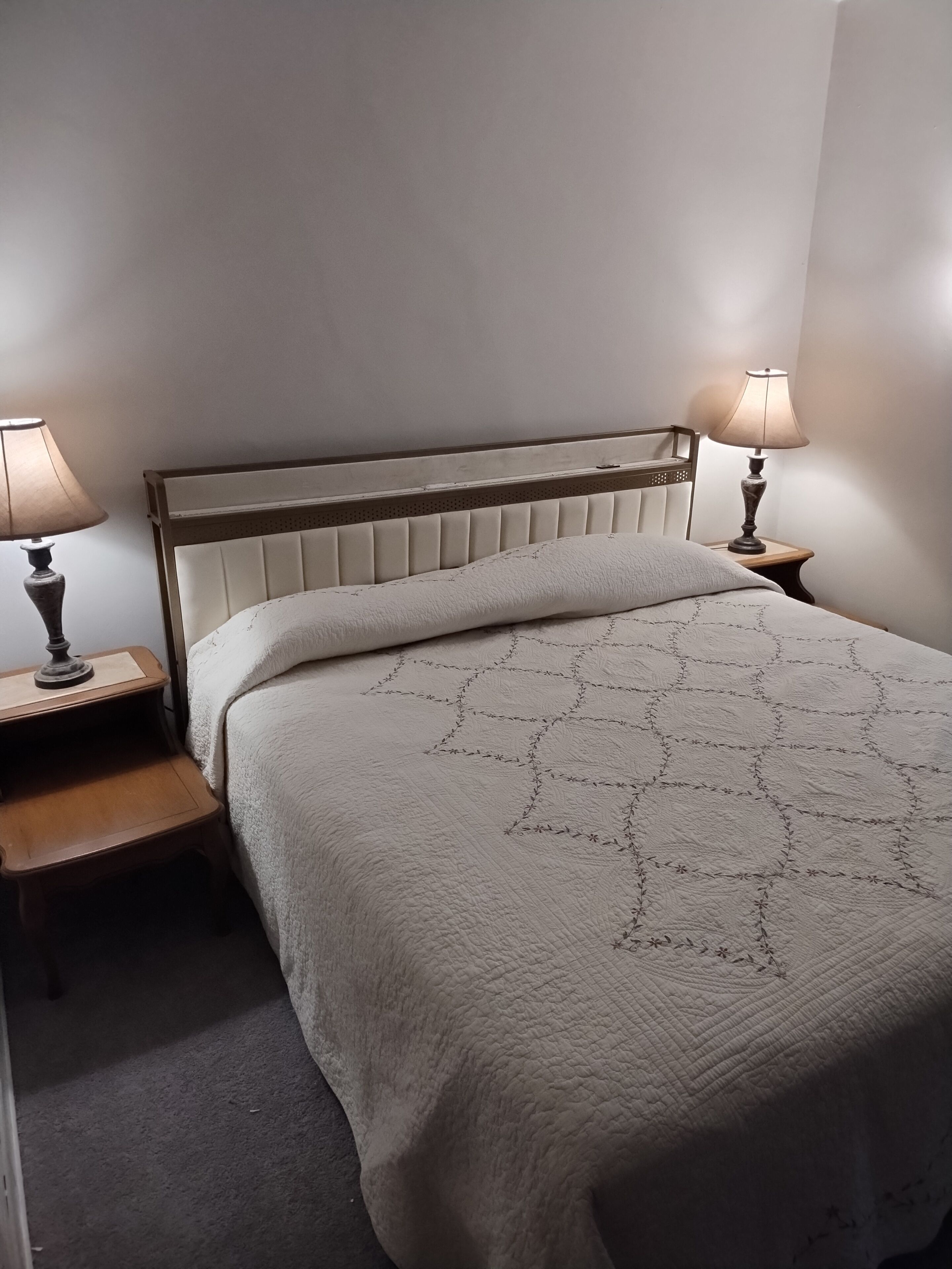 A neatly made bed with a quilted headboard, two bedside tables with lamps, and a carpeted floor.