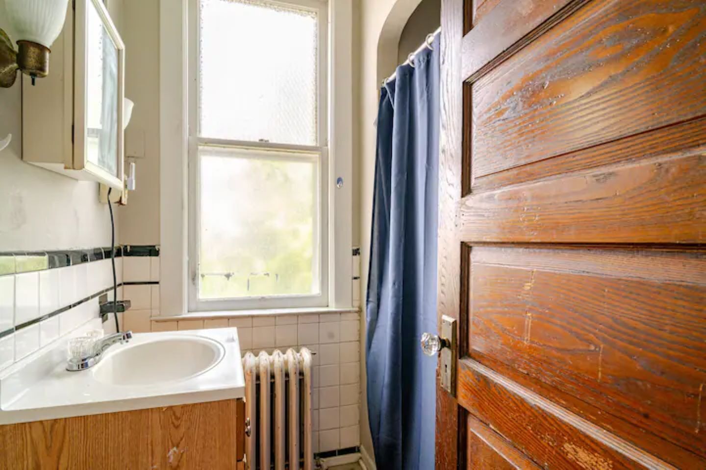 A bathroom with a wooden door, a white sink, a window with a view, and a blue shower curtain.