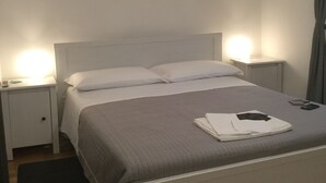 A neatly made bed with a grey blanket and white pillows, two bedside lamps, a white nightstand, and a black rug on a wooden floor.