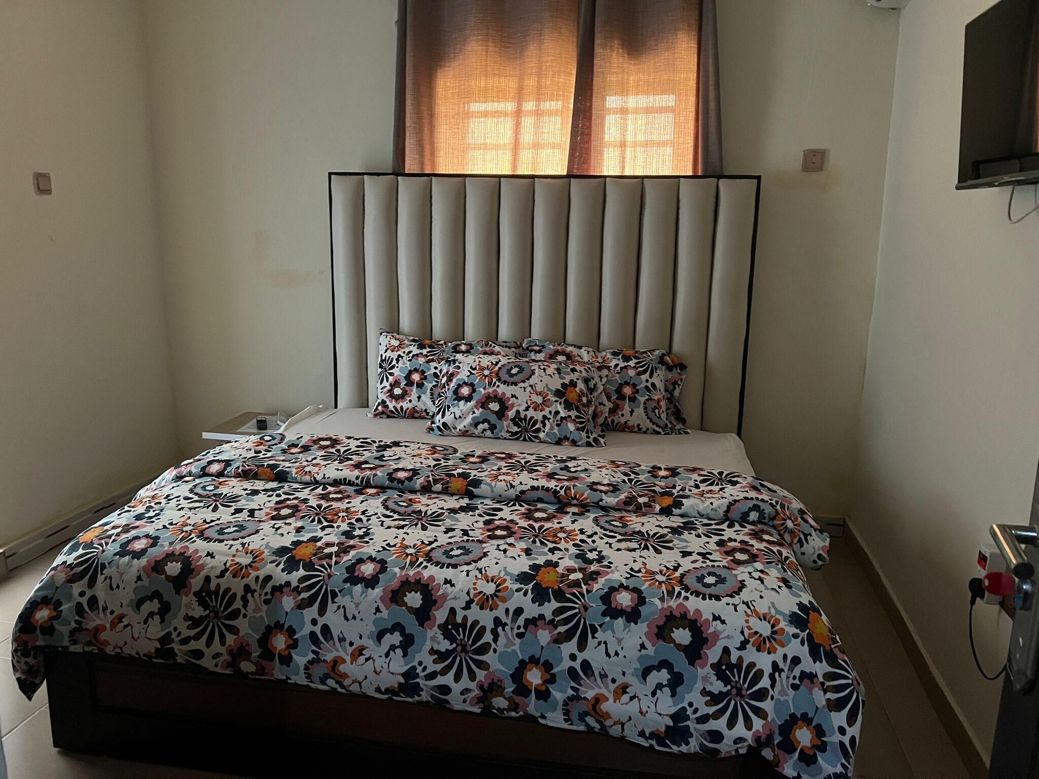 A bedroom with a bed, a headboard, a window with curtains, and a wall-mounted TV.