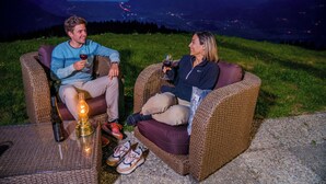 Two people seated on wicker chairs outdoors at night, holding wine glasses, with a bottle and a lit candle on a table. - Mazot Douillet de Montagne Isolé en Pleine Nature en Autonomie D'énergie (Les Giettes)